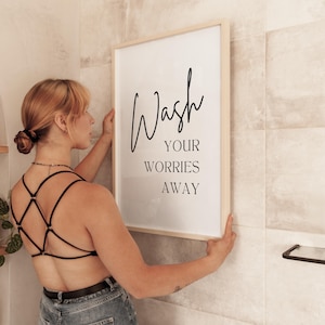 May include: A white framed print with black text that reads "Wash your worries away" hanging on a tiled wall. A person is hanging the print on the wall.