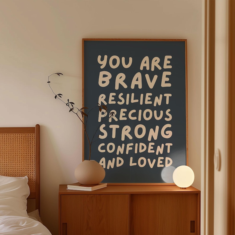 May include: A framed poster with the words "YOU ARE BRAVE RESILIENT PRECIOUS STRONG CONFIDENT AND LOVED" in white lettering on a dark blue background. The poster is displayed above a wooden cabinet with a vase and a small lamp.