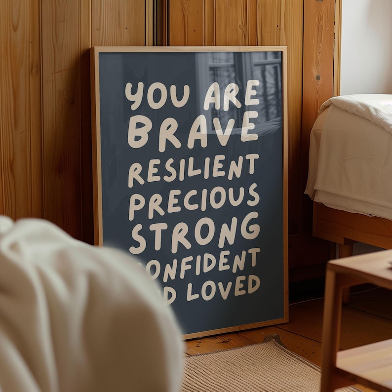 May include: A framed poster with a navy blue background and white text. The text reads "YOU ARE BRAVE RESILIENT PRECIOUS STRONG CONFIDENT LOVED". The frame is a light brown color. The poster is leaning against a wooden wall.