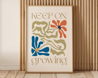 Op de afbeelding: Een ingelijste kunstdruk met de woorden "KEEP ON growing" in een beige lettertype. Het kunstwerk toont abstracte bloemmotieven in blauw, oranje en groen op een crèmekleurige achtergrond. De lijst is lichtbruin en de print wordt tegen een houten muur getoond.