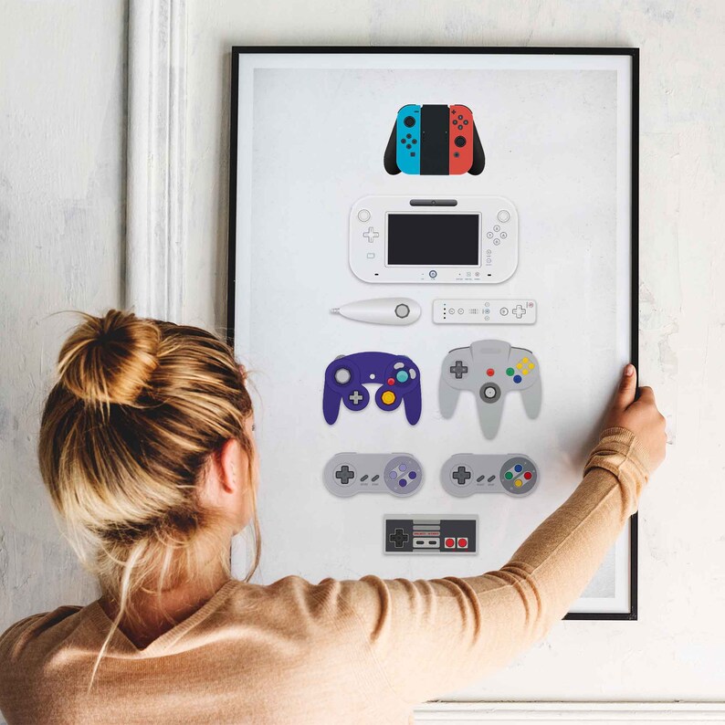 May include: A framed print featuring a collection of video game controllers and consoles, including a Nintendo Switch, a Wii, a GameCube, a Super Nintendo, and a Nintendo Entertainment System. The print is on a white background with black borders.