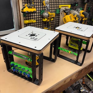 May include: Two black and white router tables with green and blue tool holders. The tables have a white surface with a black frame and a wooden edge. Power tools are mounted on the wall in the background.