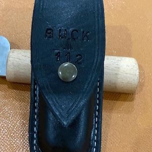 May include: A black leather sheath featuring a pointed flap with a metal snap closure. The text BUCK 112 is embossed on the front flap. The sheath has visible white stitching along the edges and is designed to hold a folding knife securely.
