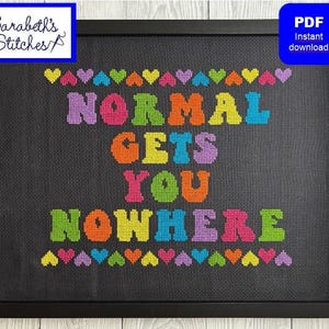 May include: Framed cross-stitch art with the text "NORMAL GETS YOU NOWHERE" in colorful letters. The design includes rows of hearts above and below the text. The artwork is framed in a black frame and includes a "PDF Instant download" icon.