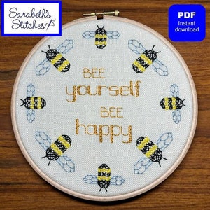May include: A white embroidery hoop with a beige background featuring a cross-stitch design of bees and the text "Bee yourself, Bee happy".
