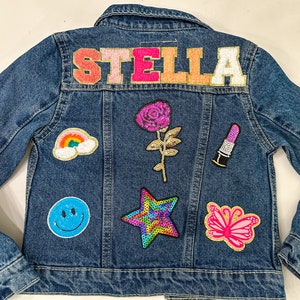 May include: A blue denim jacket with the name "STELLA" in pink, white, and orange letters on the back. The jacket is decorated with colorful patches, including a rainbow, a smiley face, a star, a rose, a butterfly, and a lipstick.