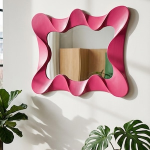 May include: A rectangular wall mirror featuring a bold, wavy magenta frame with a continuous, fluid design. The mirror surface is clear and reflective, capturing the surrounding space. The frame has a smooth, matte finish and a distinct, undulating shape.
