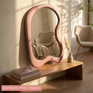 May include: A large, floor-standing mirror featuring an irregular, organic, wavy shape with a smooth, matte antique pink frame. The reflective surface shows a room interior with a chair and a blanket. The mirror rests on a wooden surface.