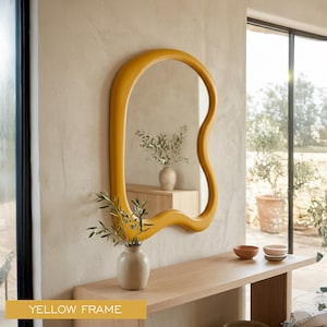 May include: A wall-mounted mirror featuring a thick, smooth, mustard yellow frame with an irregular, organic, wavy shape. The mirror reflects a room interior with a light-colored wall and a small vase containing green leafy branches.