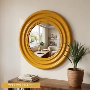 May include: A circular wall mirror featuring a thick, smooth, mustard yellow frame with concentric ridged detailing. The reflective glass surface shows a room interior with a desk, a window, and a potted plant. The text YELLOW FRAME is visible.