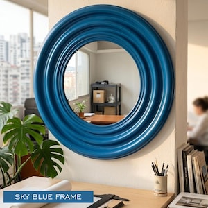 May include: A circular wall mirror featuring a glossy, deep blue frame with concentric, rippled circular ridges. The mirror reflects an interior room with a window, a shelving unit, and a desk. The frame has a smooth, reflective finish.