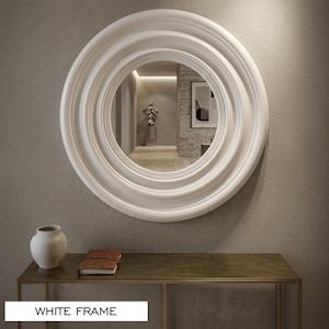 May include: A large circular wall mirror featuring a thick, multi-layered white frame with concentric moulded rings. The mirror surface reflects an interior space. The text "WHITE FRAME" is displayed in the lower left corner of the image.