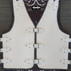 以下が含まれることがあります： 白の革ベスト。テクスチャード加工の模様、濃いグレーのトリム、シルバーカラーのバックルが特徴です。装飾的なスタッドとペイズリー柄の裏地が付いています。「LEATHER ESCORT」のブランドロゴが内側に表示されています。