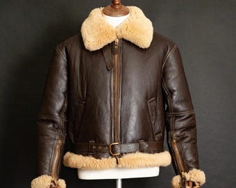 May include: A brown leather jacket with a tan shearling collar and lining. The jacket features a front zipper, a waist belt with a buckle, and zippered cuffs. The jacket is displayed on a mannequin against a dark background.