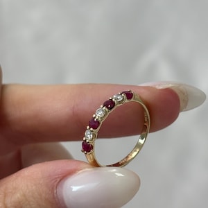 May include: A gold ring featuring alternating red gemstones and clear diamonds. The ring is held up, showcasing its design and the sparkle of the stones. The ring's band is a polished gold color.