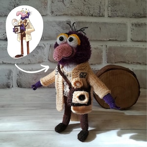 May include: A crocheted doll of a purple creature with a large pink nose, wearing a beige knit jacket, a patterned sweater, and brown trousers. The doll has purple hands, dark brown shoes, and a small brown camera bag slung across its shoulder.