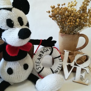 May include: A white and black knitted Mickey Mouse doll wearing a red bow tie and a sailor hat, holding a white knitted life preserver with the text "STEAMBOAT WILLIE" in black. The doll is sitting on a wooden box with a white wooden "LOVE" sign in the background.