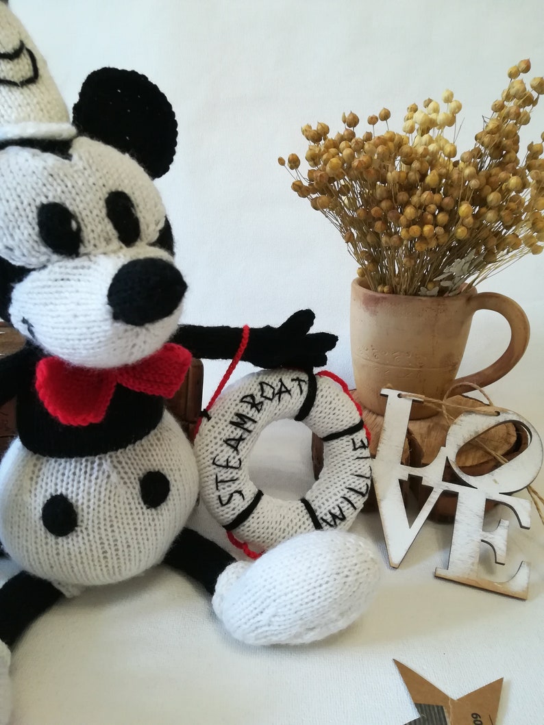 May include: A white and black knitted Mickey Mouse doll wearing a red bow tie and a sailor hat, holding a white knitted life preserver with the text "STEAMBOAT WILLIE" in black. The doll is sitting on a wooden box with a white wooden "LOVE" sign in the background.
