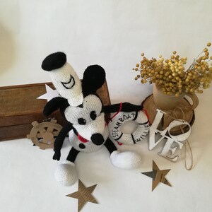 May include: A white and black knitted Mickey Mouse toy with a red and white life preserver that says "STEAMBOAT WILLIE". The toy is sitting on a white surface with a wooden box, a wooden sign that says "LOVE", and a bouquet of dried yellow flowers.