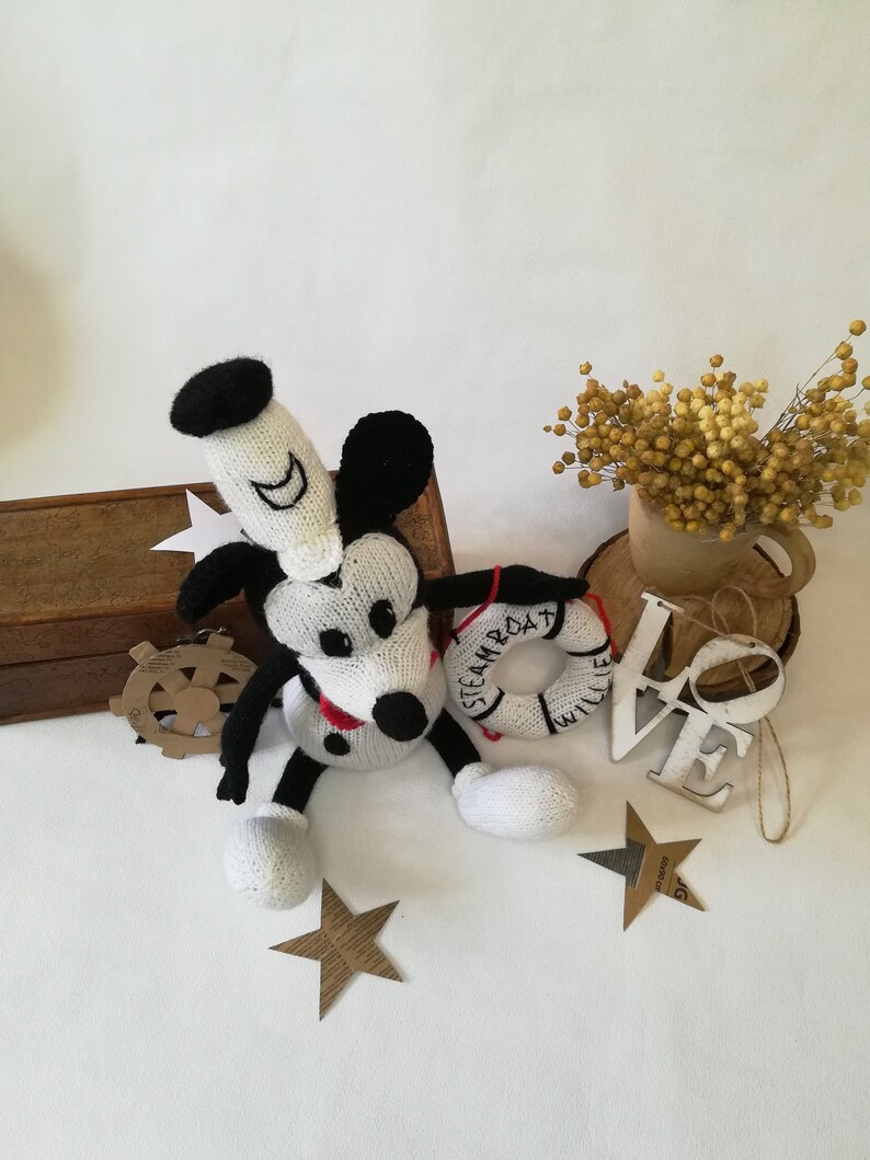 May include: A white and black knitted Mickey Mouse toy with a red and white life preserver that says "STEAMBOAT WILLIE". The toy is sitting on a white surface with a wooden box, a wooden sign that says "LOVE", and a bouquet of dried yellow flowers.
