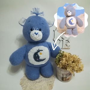 以下が含まれることがあります： 胸に白い三日月と星がついた青いニットのケアベア。クマは手にハートを持っています。クマは白いドイリーの上に座っています。