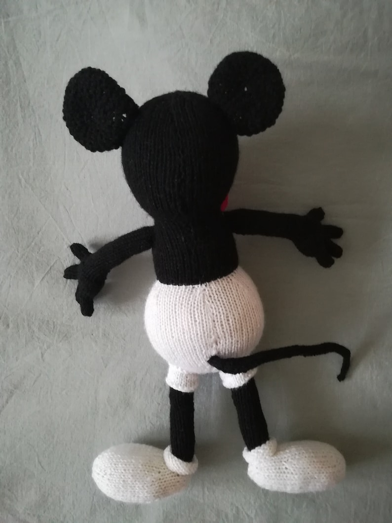 May include: A black and white knitted Mickey Mouse toy with a red nose. The toy is lying on its back with its arms outstretched and its legs bent.