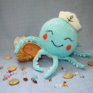 May include: A knitted blue octopus wearing a white sailor hat with an anchor embroidered on it. The octopus has white pearl beads around its tentacles and is sitting on a seashell. There are small colorful beads scattered around the octopus.