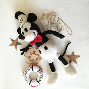 May include: A white and black knitted Mickey Mouse toy with a red bow tie, holding a wooden ship's wheel. The toy is surrounded by a life preserver with the text "Steamboat Willie", a wooden star, and a wooden sign that says "Love".