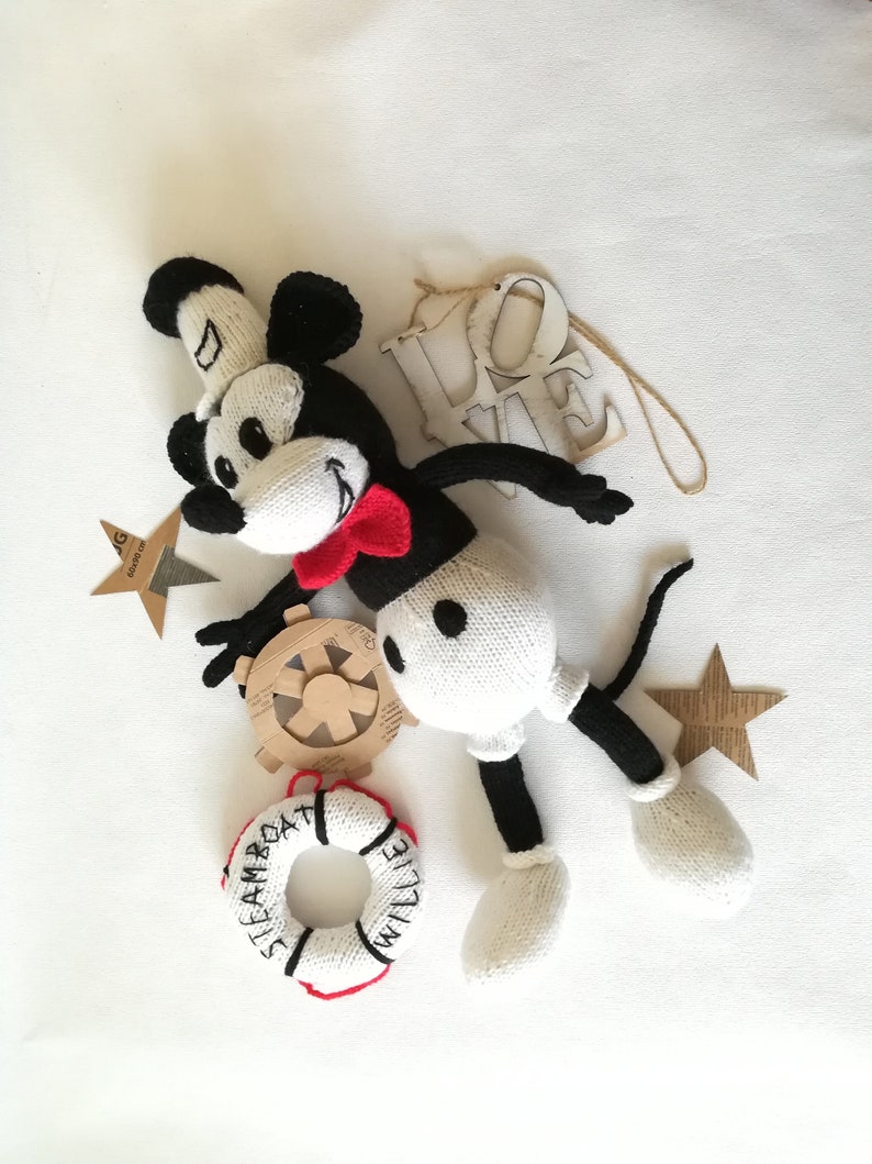 May include: A white and black knitted Mickey Mouse toy with a red bow tie, holding a wooden ship's wheel. The toy is surrounded by a life preserver with the text "Steamboat Willie", a wooden star, and a wooden sign that says "Love".
