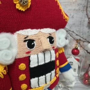 Puede incluir: Un mu&ntilde;eco de cascanueces de punto con un gorro rojo, barba blanca y una boca a rayas blancas y negras. El mu&ntilde;eco lleva un uniforme rojo y azul con botones dorados.