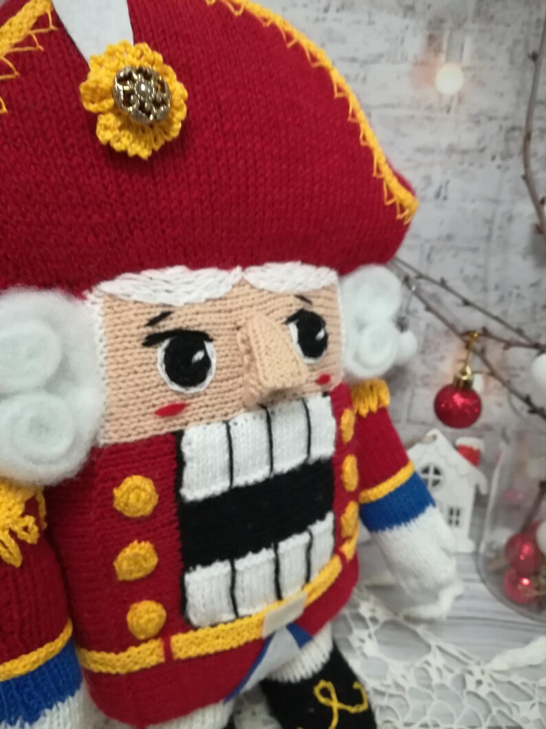 Puede incluir: Un mu&ntilde;eco de cascanueces de punto con un gorro rojo, barba blanca y una boca a rayas blancas y negras. El mu&ntilde;eco lleva un uniforme rojo y azul con botones dorados.