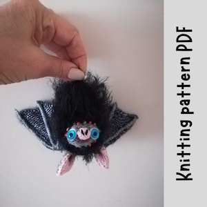 Può includere: Giocattolo amigurumi a forma di pipistrello lavorato a maglia con pelliccia nera, ali grigie, orecchie rosa e occhi blu. Il pipistrello ha perline colorate intorno al collo.