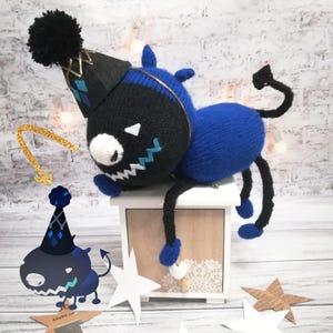 May include: A blue and black knitted plush toy with a party hat and a tail. The toy is sitting on a small wooden box with a white top.