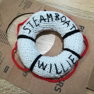 May include: White knitted life preserver with black and red trim. The words "STEAMBOAT WILLIE" are embroidered on the front.