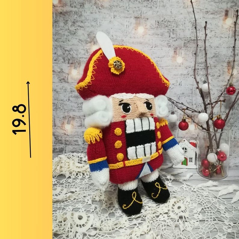 Puede incluir: Un mu&ntilde;eco de cascanueces de punto que lleva un gorro rojo y blanco con ribete amarillo y azul. El mu&ntilde;eco mide aproximadamente 50 cm de alto y tiene barba y bigote blancos.