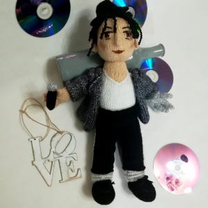 Puede incluir: Muñeco de punto de una persona que lleva una chaqueta gris, una camisa blanca, pantalones negros y zapatos negros. El muñeco sostiene un micrófono en una mano y un guante plateado en la otra. El muñeco está rodeado de CDs y un letrero de madera que dice "LOVE".