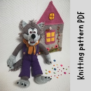 Könnte beinhalten: Gestrickte graue Wolfspuppe, die eine lila Latzhose und einen orangefarbenen Schal trägt. Der Wolf sitzt auf einer weißen Oberfläche mit einem rosa gestrickten Haus im Hintergrund. Der Text "Knitting pattern PDF" befindet sich auf der rechten Seite des Bildes.
