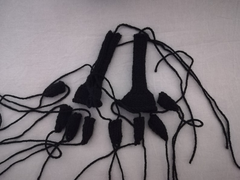 May include: A set of black knitted fingerless gloves with long strings attached. The gloves are laid out on a white surface.