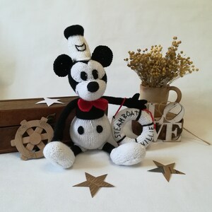 May include: A white and black knitted Mickey Mouse toy wearing a sailor hat and holding a red and white life preserver with the text "Steamboat Willie" on it.