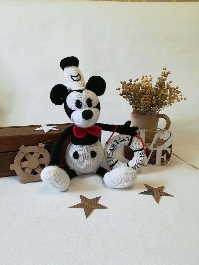 May include: A white and black knitted Mickey Mouse toy wearing a sailor hat and holding a red and white life preserver with the text "Steamboat Willie" on it.