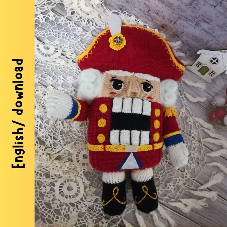 Puede incluir: Un mu&ntilde;eco de cascanueces de punto con un gorro rojo, barba blanca y un uniforme amarillo y azul. El mu&ntilde;eco est&aacute; de pie sobre una superficie de crochet blanco.