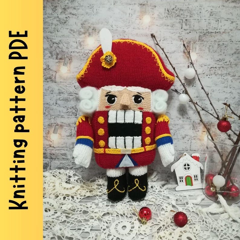 Puede incluir: Mu&ntilde;eco cascanueces de punto con un gorro rojo, barba blanca y un uniforme rojo y amarillo. El mu&ntilde;eco est&aacute; de pie sobre una superficie blanca con un fondo blanco y rojo.
