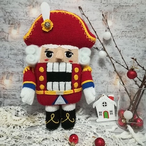 May include: A knitted nutcracker doll with a red hat, white beard, and a yellow and blue uniform. The doll is standing on a white surface with a white and red background.