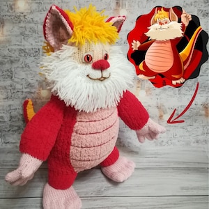 May include: A red and pink knitted plush toy with a white beard and yellow hair. The toy has a long tail and is shaped like a dragon. The toy is sitting on a wooden surface.