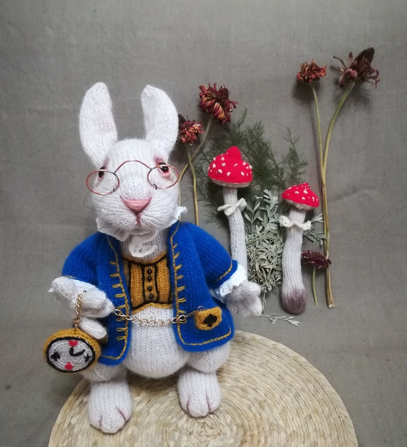 Pu&ograve; includere: Bambola di coniglio bianco fatta a mano con una giacca blu con finiture dorate, un gilet dorato e occhiali rotondi. Il coniglio tiene in mano un orologio da tasca ed &egrave; vicino a decorazioni di funghi rossi e bianchi. La bambola &egrave; su un tappetino intrecciato.