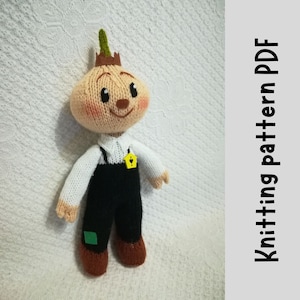 May include: Knitted amigurumi doll with a brown head shaped like an onion, wearing a white shirt, black overalls, and brown shoes. The doll has a green stem on top of its head and a yellow heart-shaped tag on its chest.