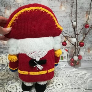 Puede incluir: Un cascanueces de juguete de punto en forma de soldado. El cascanueces lleva un uniforme rojo y blanco con un cintur&oacute;n negro y ribetes amarillos. El cascanueces tiene una barba blanca y un sombrero negro con ribetes amarillos.