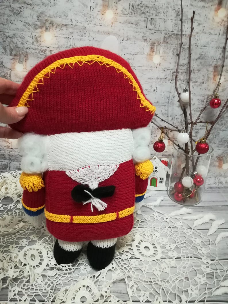Puede incluir: Un cascanueces de juguete de punto en forma de soldado. El cascanueces lleva un uniforme rojo y blanco con un cintur&oacute;n negro y ribetes amarillos. El cascanueces tiene una barba blanca y un sombrero negro con ribetes amarillos.