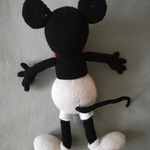 May include: A black and white knitted Mickey Mouse toy with a red bow tie. The toy is lying on its back with its arms outstretched and its legs bent. The toy has a long tail.