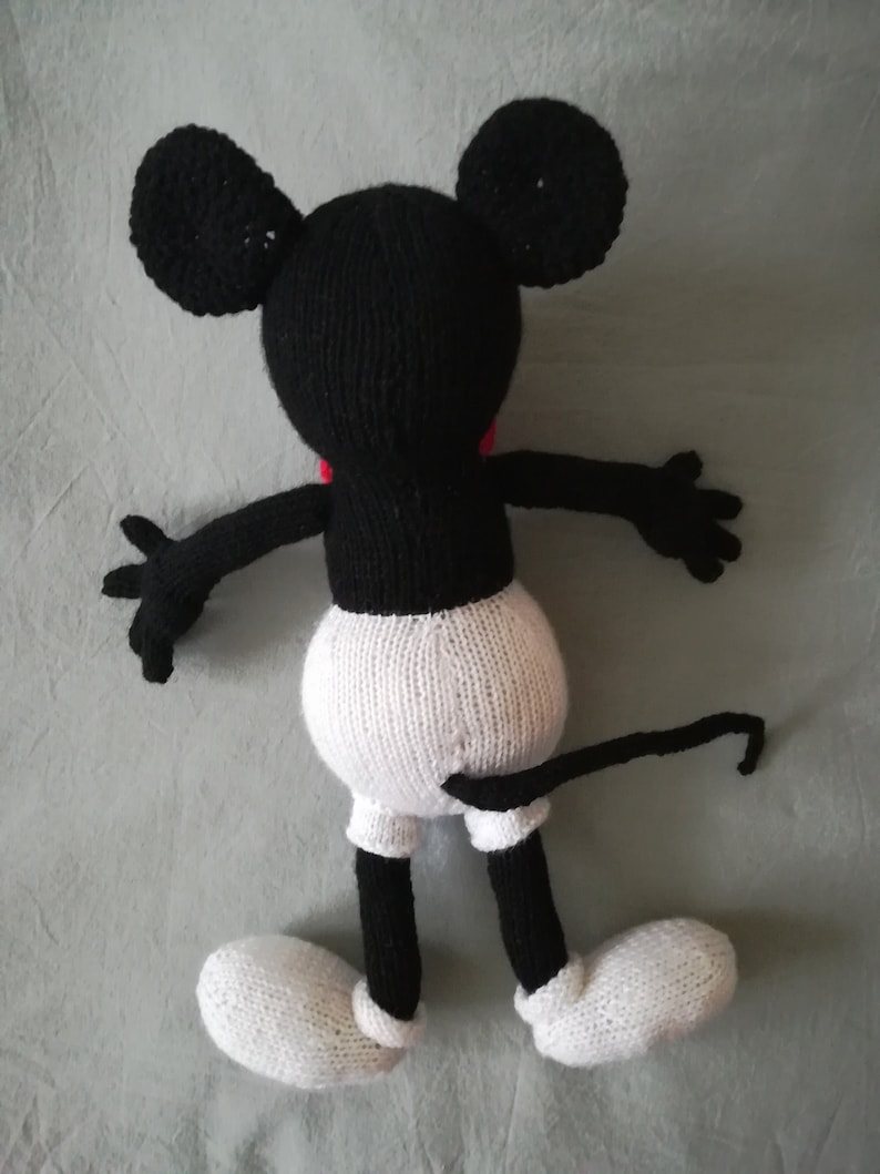 May include: A black and white knitted Mickey Mouse toy with a red bow tie. The toy is lying on its back with its arms outstretched and its legs bent. The toy has a long tail.